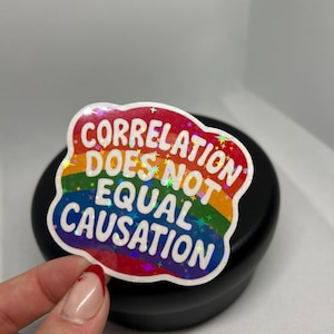 Peut inclure: Un autocollant coloré avec le texte "CORRELATION DOES NOT EQUAL CAUSATION" en blanc, encadré d'un motif arc-en-ciel. L'autocollant a un effet holographique et est tenu par une main.