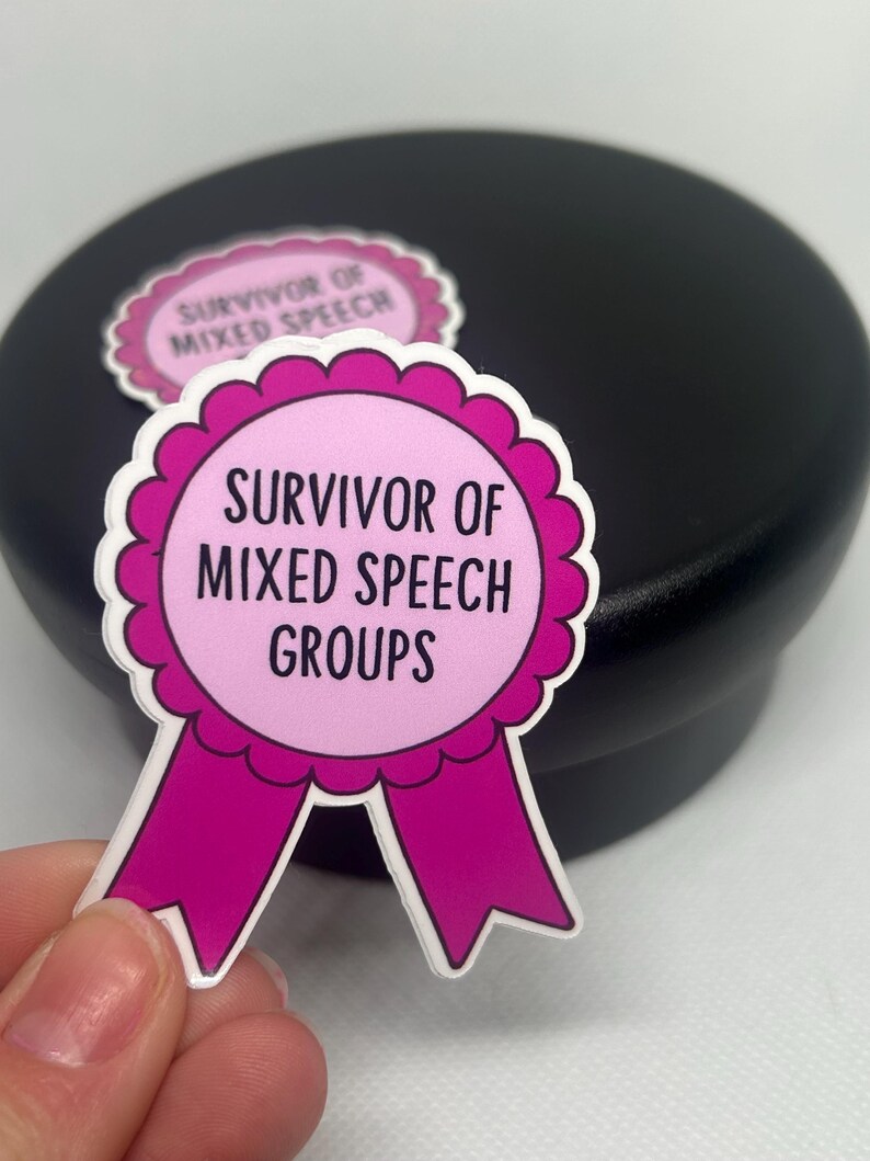 K&ouml;nnte beinhalten: Ein rosa-wei&szlig;er Aufkleber mit dem Text "Survivor of Mixed Speech Groups" in einem kreisf&ouml;rmigen Design mit einem Band. Ein weiterer Aufkleber ist im Hintergrund sichtbar. Der Aufkleber wird von einer Hand gehalten.