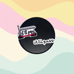 May include: Black round object with two stickers. One sticker features a street scene with a lamppost and the text "it'll pass". The other sticker has the text "it'll pass" in a cursive font. The background is a pastel color scheme.