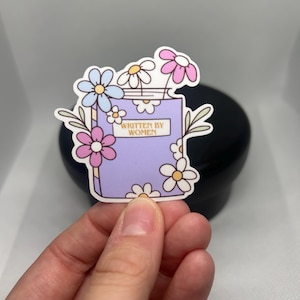 May include: A sticker featuring a purple book with the text "WRITTEN BY WOMEN" and surrounded by colorful flowers. The flowers include pink, blue, and white petals. The sticker has a white border.