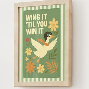 May include: A framed art print featuring a cartoon duck wearing a pilot's cap and scarf, with the text "WING IT 'TIL YOU WIN IT" on a green background. The artwork includes flowers and leaves in shades of yellow, orange, and green.