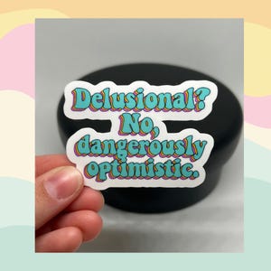 May include: A white sticker with the text "Delusional? No, dangerously optimistic." The text is in a retro font with a teal fill and a colorful outline. The sticker is held in a hand against a black background.