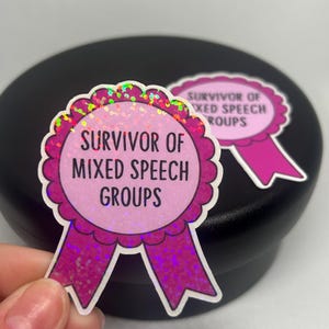 K&ouml;nnte beinhalten: Zwei rosa und glitzernde Aufkleber mit dem Text "Survivor of Mixed Speech Groups" in schwarzer Schrift. Die Aufkleber sind wie Schleifen geformt, mit einem gewellten Rand und einem rosa Band unten.