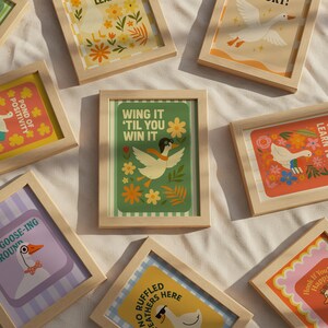 May include: A collection of framed art prints with whimsical goose illustrations. The prints feature phrases like "Wing It 'Til You Win It" and "Pond of Positivity" with floral and striped backgrounds. The frames are made of light-colored wood.