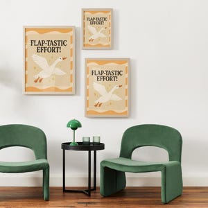 May include: Three framed art prints with the text "FLAP-TASTIC EFFORT!" and a white goose illustration. The prints are in a light wood frame. The room also features two green chairs and a black side table.