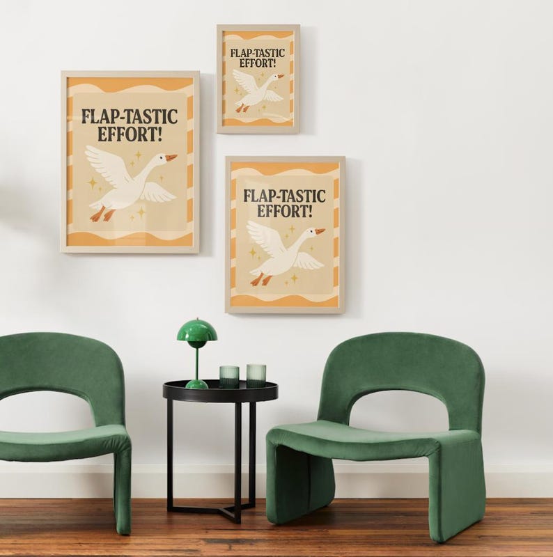 May include: Three framed art prints with the text "FLAP-TASTIC EFFORT!" and a white goose illustration. The prints are in a light wood frame. The room also features two green chairs and a black side table.