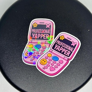 Puede incluir: Dos pegatinas rosas de teléfono plegable con el texto "PROFESSIONAL YAPPER". Las pegatinas tienen un diseño de flor y mariposa. Los botones del teléfono incluyen una carita sonriente y otras formas coloridas.