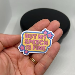 May include: A sticker with the text "BUT MY SPEECH IS FINE" in a yellow and pink design. The sticker has a white border and pink star accents. The sticker is held in a hand.