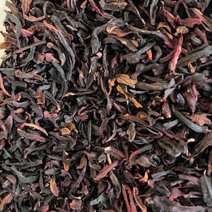 May include: Close-up of dried hibiscus flowers, displaying a mix of dark burgundy and reddish-pink shades. The petals and sepals are visible, creating a textured appearance. This image highlights the natural, organic form of the tea leaves.
