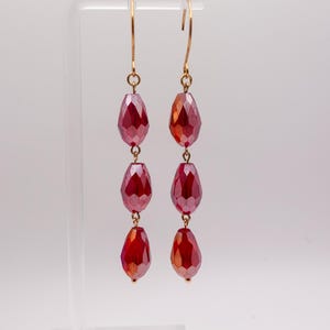 May include: A pair of dangling earrings featuring three faceted, teardrop-shaped red beads on each side. The beads are suspended from gold-toned metal hooks, creating a vertical arrangement that catches the light with its polished, reflective surfaces.