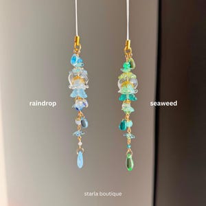 Op de afbeelding: Twee kralenhangers, &eacute;&eacute;n blauw en &eacute;&eacute;n groen, met goudkleurige accenten. De blauwe hanger is gelabeld met "raindrop" en de groene hanger met "seaweed". De hangers hebben een verscheidenheid aan kralen en vormen, waaronder bloem- en druppelvormige ontwerpen. De tekst "starla boutique" staat onderaan.