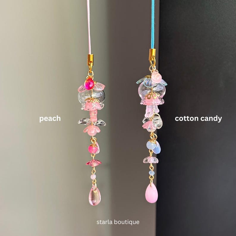 Op de afbeelding: Twee bedelhangers, &eacute;&eacute;n met het label "perzik" en de andere met "suikerspin". Elke hanger heeft een reeks roze, heldere en goudkleurige kralen, met een druppelvormige kraal aan de onderkant. De bedels hangen aan een roze en blauw koord.