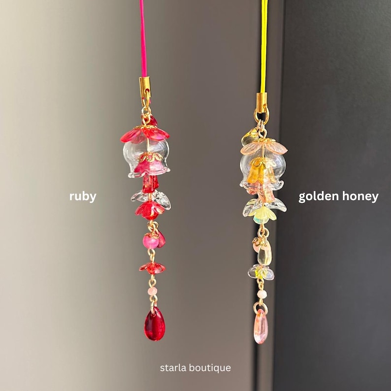 Op de afbeelding: Twee decoratieve hangende bedels. E&eacute;n is robijnkleurig met een roze koord, de andere is goudhoningkleurig met een geel koord. Beide hebben bloemen- en kralenontwerpen. De tekst "ruby", "golden honey" en "starla boutique" is zichtbaar.