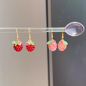 May include: Three pairs of dangle earrings with raspberry designs. The earrings have gold-coloured hooks and feature red and pink raspberry charms, green leaves, and small pink flowers. The earrings are displayed on a clear, curved rod.