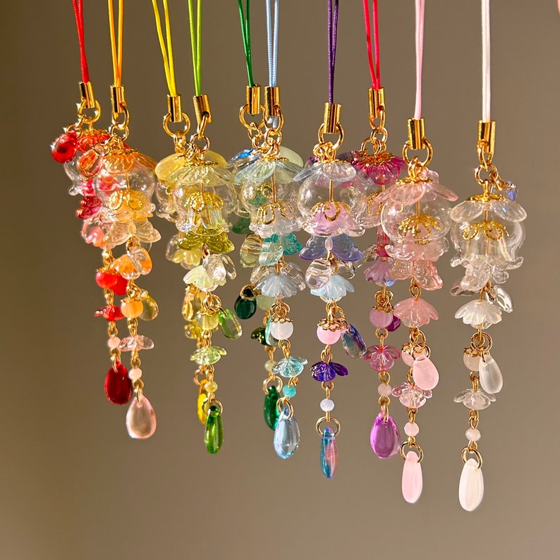 Op de afbeelding: Een verzameling kleurrijke, bungelende bedels met goudkleurige metalen accenten. Elke bedel heeft een bloemvormige bovenkant met trapsgewijze kralen en druppelvormige hangers. De bedels zijn opgehangen aan dunne, felgekleurde koorden.