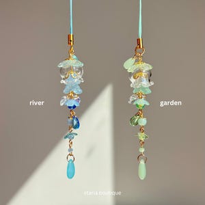 Op de afbeelding: Twee decoratieve hangende bedels. E&eacute;n is blauw en de andere is groen. De blauwe bedel heeft het woord "rivier" ernaast. De groene bedel heeft het woord "tuin" ernaast. Beide bedels hebben gouden accenten en de woorden "starla boutique" onderaan.