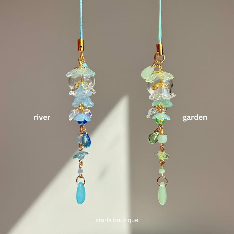 Op de afbeelding: Twee decoratieve hangende bedels. E&eacute;n is blauw en de andere is groen. De blauwe bedel heeft het woord "rivier" ernaast. De groene bedel heeft het woord "tuin" ernaast. Beide bedels hebben gouden accenten en de woorden "starla boutique" onderaan.