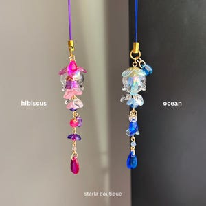Op de afbeelding: Twee bedelhangers met kralen, &eacute;&eacute;n met het label "hibiscus" met roze en paarse kralen, en de andere met het label "ocean" met blauwe kralen. Beide bedels hebben goudkleurige metalen accenten en zijn opgehangen aan dunne koorden. De tekst "starla boutique" staat onderaan.