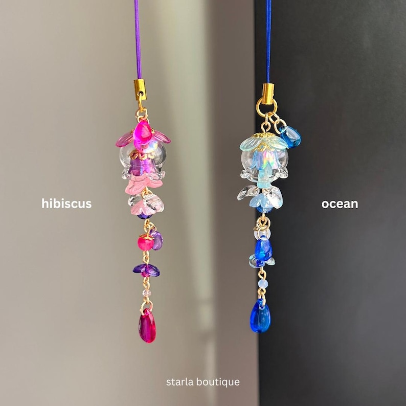 Op de afbeelding: Twee bedelhangers met kralen, &eacute;&eacute;n met het label "hibiscus" met roze en paarse kralen, en de andere met het label "ocean" met blauwe kralen. Beide bedels hebben goudkleurige metalen accenten en zijn opgehangen aan dunne koorden. De tekst "starla boutique" staat onderaan.