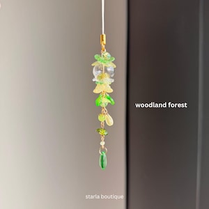 Op de afbeelding: Een delicate, groen en gouden bedel met een bos thema. De bedel heeft een heldere glazen bol, groene en gele bloemvormige kralen en kleine groene kralen. De tekst "woodland forest" en "starla boutique" zijn zichtbaar.