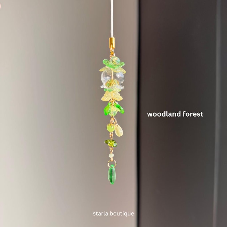 Op de afbeelding: Een delicate, groen en gouden bedel met een bos thema. De bedel heeft een heldere glazen bol, groene en gele bloemvormige kralen en kleine groene kralen. De tekst "woodland forest" en "starla boutique" zijn zichtbaar.