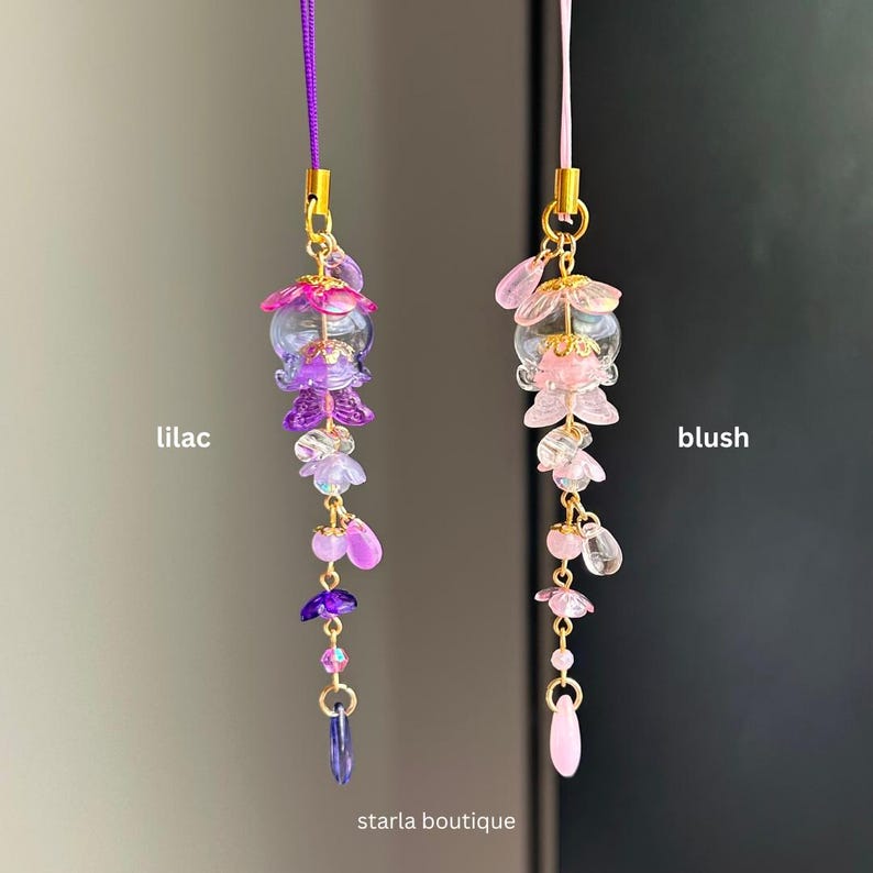 Op de afbeelding: Twee decoratieve bloemenbedels, &eacute;&eacute;n in lila en de andere in blush, hangen aan paarse en roze koorden. De bedels hebben gouden accenten en zijn gelabeld met de kleuren. De tekst "starla boutique" staat onderaan.