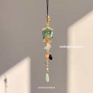 Op de afbeelding: Een decoratieve bedel met een goudkleurige sluiting en een zwart koord. De bedel heeft een cluster van kleurrijke kralen en glaselementen in tinten groen, oranje en zwart, met de tekst "midnight autumn" en "starla boutique".