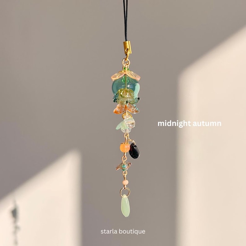 Op de afbeelding: Een decoratieve bedel met een goudkleurige sluiting en een zwart koord. De bedel heeft een cluster van kleurrijke kralen en glaselementen in tinten groen, oranje en zwart, met de tekst "midnight autumn" en "starla boutique".