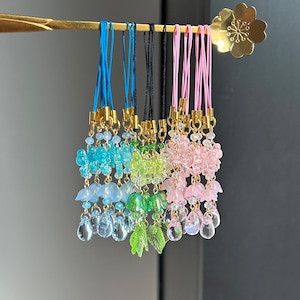 Op de afbeelding: Een set van negen kleurrijke telefooncharms met verschillende ontwerpen. Elke charm heeft een bloemvormige bovenkant met een hangende kristal-achtige kraal. De charms zijn bevestigd aan een gouden ring en een touwtje.