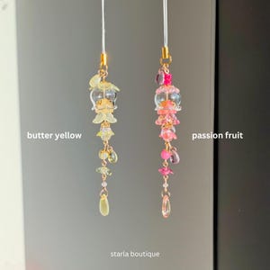 Op de afbeelding: Twee kralen telefoonbedels, &eacute;&eacute;n botergeel en &eacute;&eacute;n passievrucht. De botergele bedel heeft gele en heldere kralen met gouden accenten. De passievruchtbedel heeft roze en heldere kralen met gouden accenten. De bedels hangen aan witte koorden.