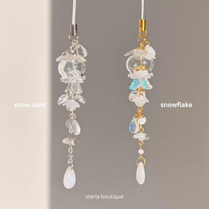 Op de afbeelding: Twee decoratieve telefoonbedels. E&eacute;n is gelabeld "snow dust" en de andere "snowflake". Beide bedels hebben heldere, bloemvormige kralen en druppelvormige hangers. De "snowflake" bedel heeft gouden accenten en iriserende kralen. De tekst "starla boutique" staat onderaan.