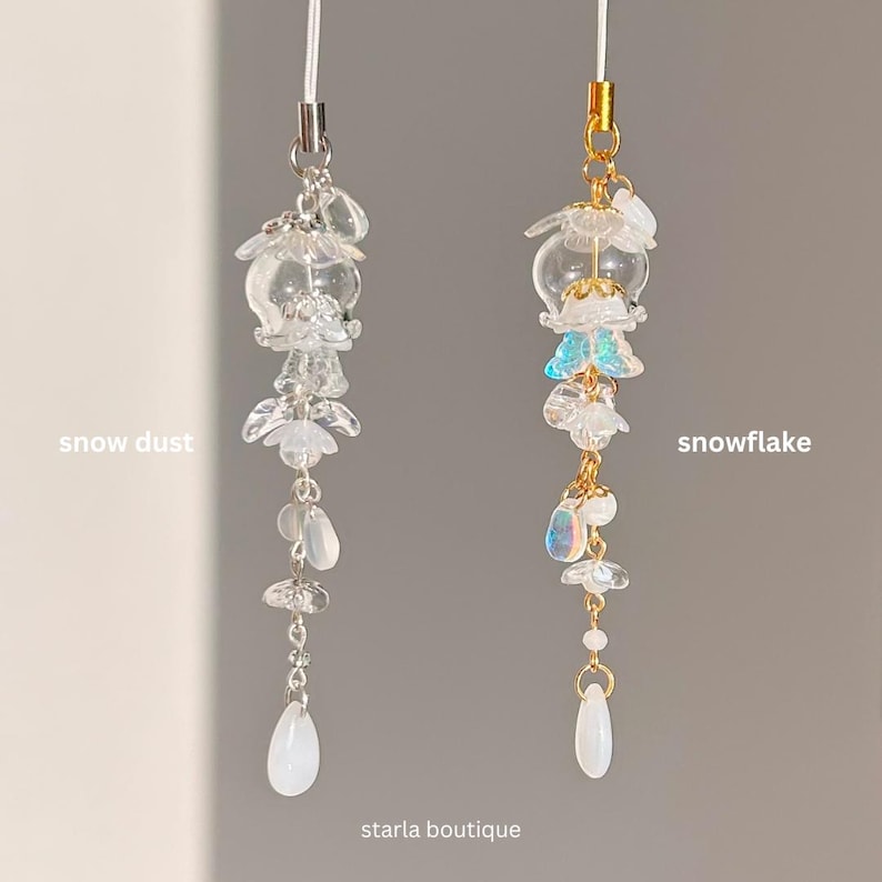 Op de afbeelding: Twee decoratieve telefoonbedels. E&eacute;n is gelabeld "snow dust" en de andere "snowflake". Beide bedels hebben heldere, bloemvormige kralen en druppelvormige hangers. De "snowflake" bedel heeft gouden accenten en iriserende kralen. De tekst "starla boutique" staat onderaan.
