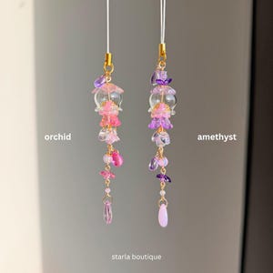 Op de afbeelding: Twee kralen telefoonbedels, &eacute;&eacute;n met het label "orchidee" en de andere "amethist". De orchidee&euml;nbedel heeft roze en heldere kralen, terwijl de amethistbedel paarse en heldere kralen heeft. Beide hebben goudkleurige accenten en een bungelende druppelkraal. De tekst "starla boutique" staat onderaan.