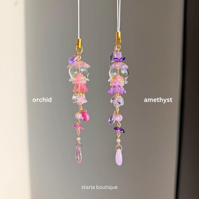 Op de afbeelding: Twee kralen telefoonbedels, &eacute;&eacute;n met het label "orchidee" en de andere "amethist". De orchidee&euml;nbedel heeft roze en heldere kralen, terwijl de amethistbedel paarse en heldere kralen heeft. Beide hebben goudkleurige accenten en een bungelende druppelkraal. De tekst "starla boutique" staat onderaan.