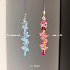 Op de afbeelding: Twee kralen telefoonbedels, &eacute;&eacute;n blauw en &eacute;&eacute;n roze. De blauwe bedel, met het label "forget me not", heeft blauwe bloem- en druppelkralen. De roze bedel, met het label "rose garden", heeft roze bloem- en druppelkralen. De winkelnaam "starla boutique" staat onderaan.