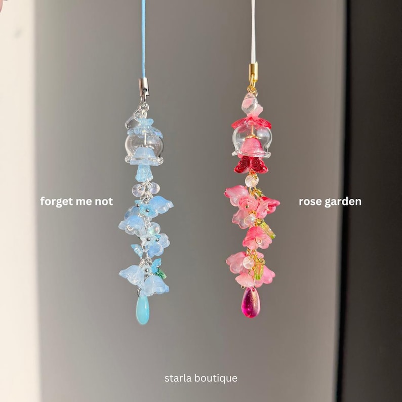 Op de afbeelding: Twee kralen telefoonbedels, &eacute;&eacute;n blauw en &eacute;&eacute;n roze. De blauwe bedel, met het label "forget me not", heeft blauwe bloem- en druppelkralen. De roze bedel, met het label "rose garden", heeft roze bloem- en druppelkralen. De winkelnaam "starla boutique" staat onderaan.