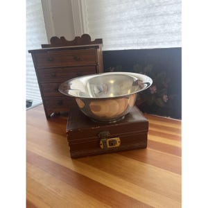May include: A shiny, silver metal bowl with a wide rim, resting on a brown, rectangular box with a gold clasp. The bowl reflects the surrounding environment. A small wooden dresser is in the background.