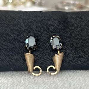 May include: A pair of gold-toned earrings with black oval-shaped gemstones. The earrings feature a cone-shaped design below the gemstone and a curved, open loop at the bottom. The earrings are displayed on a black surface.