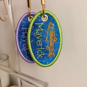 May include: Two oval embroidered patches with gold-tone metal carabiners. One patch has a lime green border, blue speckled background, orange fish outline, and the name Maverick. The other has a purple border, blue background, and the name Hayes.