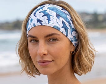 May include: A white fabric headband featuring a repeating pattern of various blue sharks and marine life illustrations. The headband is worn around the head, displaying a detailed print of different shark species in shades of blue.