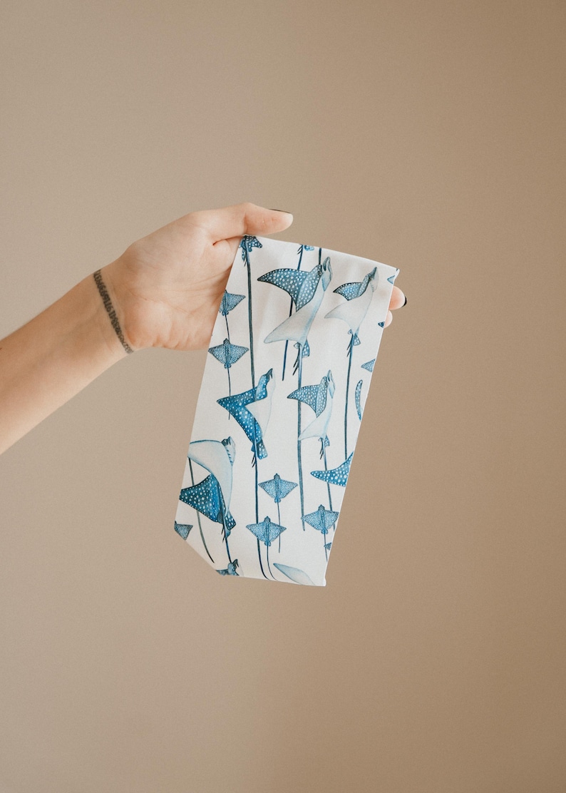 May include: A white fabric headband featuring a repeating pattern of blue spotted stingrays and manta rays. The marine life illustrations are rendered in shades of blue and white, arranged in a vertical orientation across the surface of the band.