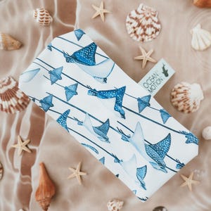 May include: A rectangular white fabric tissue box cover featuring a repeating pattern of blue spotted stingrays swimming along thin dark blue horizontal lines. A small rectangular tag attached to the side displays the text OCEAN MOTION.