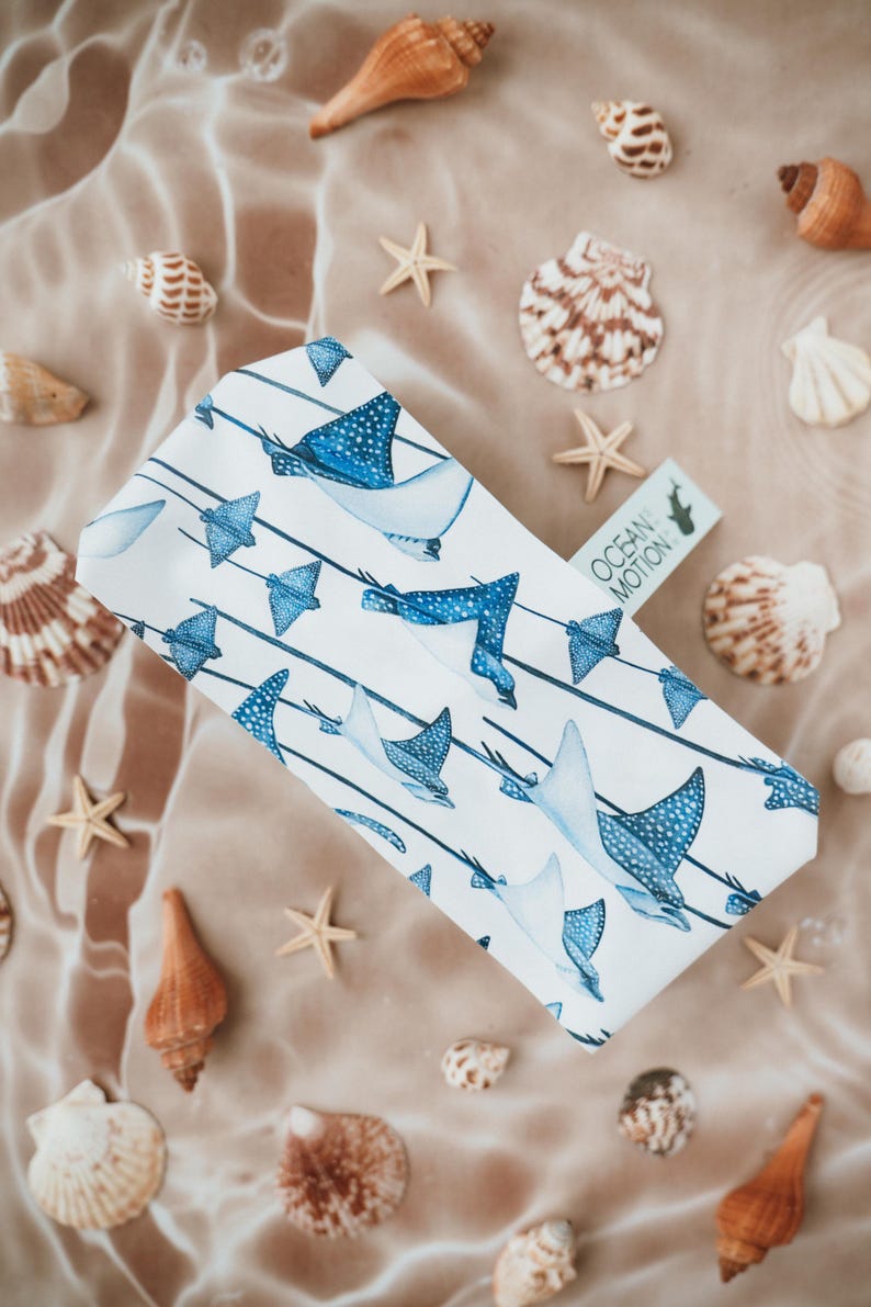 May include: A rectangular white fabric tissue box cover featuring a repeating pattern of blue spotted stingrays swimming along thin dark blue horizontal lines. A small rectangular tag attached to the side displays the text OCEAN MOTION.