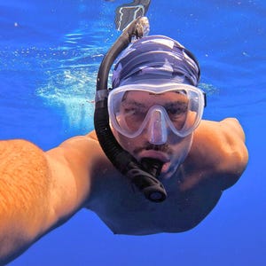 May include: A person swimming underwater wearing a clear silicone diving mask with a black frame and a black snorkel attached to the side. The person is also wearing a dark blue patterned swim cap while submerged in bright blue ocean water.