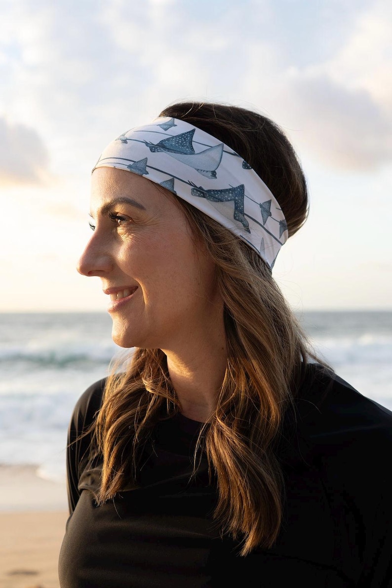 May include: A white fabric headband featuring a repeating pattern of blue and grey stingrays swimming across thin horizontal lines. The accessory is wide and fits snugly around the head, displaying a detailed marine life print on the material.