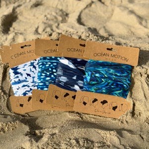 May include: Four tubular fabric headbands labeled Ocean Motion Wear on brown cardboard packaging. The patterns include a white shark print, a blue whale shark dot pattern, a dark blue sea turtle print, and a blue and green iridescent wave design.