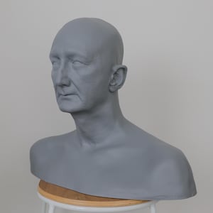 Pode incluir: Um busto de gesso cinza de uma pessoa com a cabeça careca. O busto está sobre um pedestal de madeira.