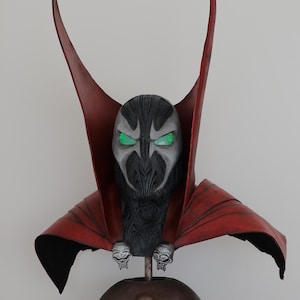 May include: A detailed bust of a character featuring a black textured mask with glowing green eyes, a high-collared red cape, and two silver skull-shaped clasps at the shoulders. The figure is mounted on a dark, rounded base with a skull emblem.