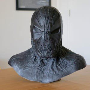 Puede incluir: Una máscara y un busto negros esculpidos, con una superficie texturizada y detallada. La máscara tiene un diseño estilizado y angular con aberturas para los ojos y patrones intrincados. El busto se extiende hasta los hombros, con un acabado texturizado similar.