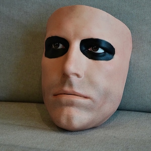 May include: A pale mask with black eye makeup, resembling a human face. The mask features a bald head, brown eyes, and a neutral expression. It is likely designed for costume or theatrical purposes.